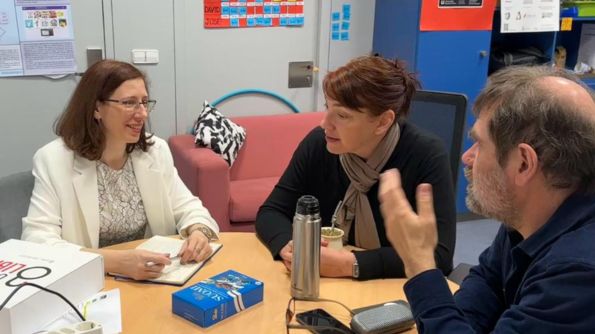 Imagen de tres personas sentadas alrededor de una mesa en una oficina. La persona a la izquierda lleva una chaqueta blanca y está escribiendo en un cuaderno. La persona en el centro sostiene una taza y tiene un termo sobre la mesa. La persona a la derecha está gesticulando con una mano mientras habla. Sobre la mesa hay varios objetos, incluyendo una caja de bombones, un par de gafas y algunos dispositivos electrónicos. En el fondo, hay pósteres, gráficos y estanterías con varios objetos.